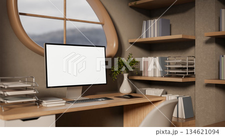 Blank screen computer on wooden study table aside bookshelf under oval window in modern living room. 134621094