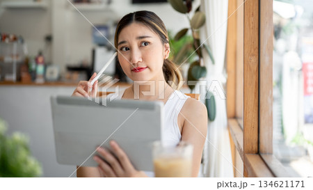 Asian woman holding stylus touching head thinking while looking away from tablet at cafe's table. 134621171