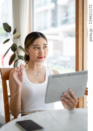 Asian woman holding stylus looking away from tablet sitting at round table beside cafe's glass wall Asian woman holding stylus looking away from tablet sitting at round table beside cafe's glass wall 134621172