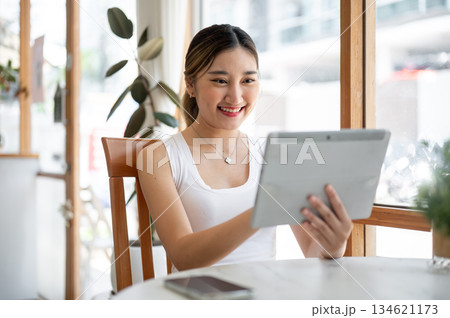 Asian woman holding and looking at the tablet while sitting at round table beside glass wall in cafe Asian woman holding and looking at the tablet while sitting at round table beside glass wall in cafe 134621173