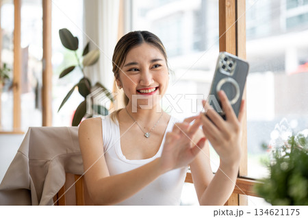 Pretty asian woman holding smartphone taking photo or selfie at table beside glass wall in the cafe. 134621175