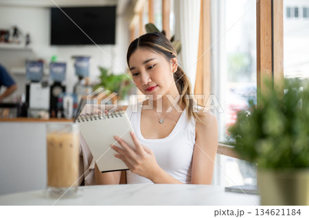 Asian woman holding notebook looking or reading with smile as sitting with ice coffee at cafe table. 134621184