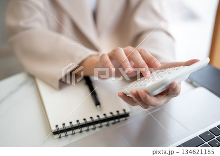 Close up of hands holding and using or pressing calculator over notebook and laptop on table in cafe 134621185