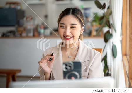 Asian woman holding pen and phone talking on video call sitting in front of a laptop at cafe's table 134621189