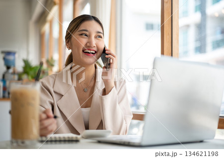 Asian woman talking on phone with pen over notebook sitting with laptop and coffee at cafe's table 134621198