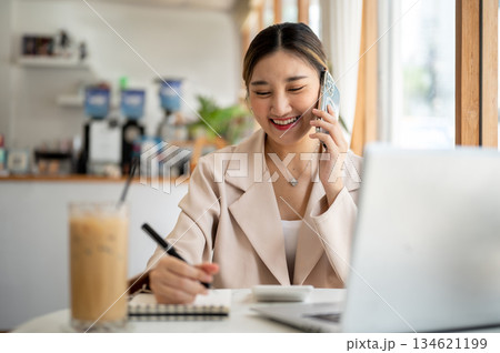 Asian woman talking on phone with pen on notebook as sitting with laptop and coffee at cafe's table. 134621199