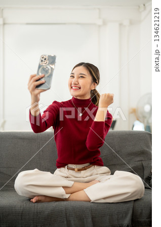 Happy asian woman looking at smartphone and making a fist while sitting on sofa in a living room. Happy asian woman looking at smartphone and making a fist while sitting on sofa in a living room. 134621219