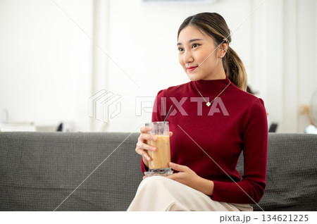 Asian woman smiling as holding ice coffee and sitting cross legged on sofa in living room or lounge 134621225