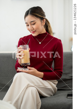 Asian woman enjoy drinking iced coffee while sitting cross legged on sofa in a living room or lounge 134621226