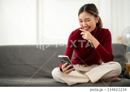 Pretty asian woman laughing as looking at phone while sitting cross legged on sofa in a living room. 134621234