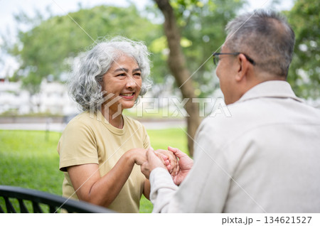 Close up of an old woman smiling at old man while holding hand and sitting together on bench in park 134621527