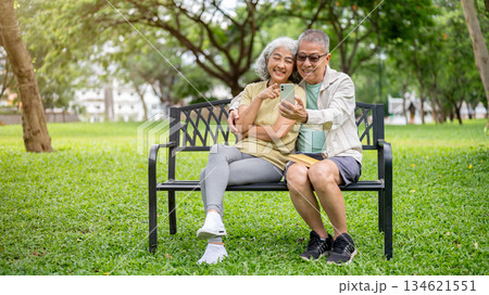 Glasses old man holding phone taking photo watching video with woman while sitting on bench in park. Glasses old man holding phone taking photo watching video with woman while sitting on bench in park. 134621551