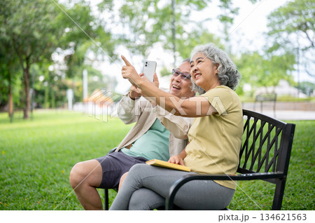 Glasses old man holding phone taking pictures where woman pointing sitting together on bench in park Glasses old man holding phone taking pictures where woman pointing sitting together on bench in park 134621563