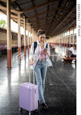 Asian woman looking at phone and holding a purple luggage while standing or waiting at train station. Asian woman looking at phone and holding a purple luggage while standing or waiting at train station. 134621669