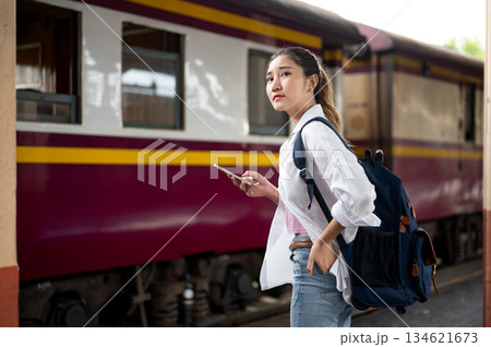 Asian woman with backpack holding phone and looking ahead while standing or waiting at train station Asian woman with backpack holding phone and looking ahead while standing or waiting at train station 134621673