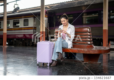 Pretty asian woman holding and looking at smartphone while sitting on wooden bench at train station. 134621687