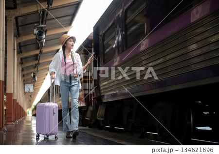 Asian woman holding phone looking at purple carriage and dragging luggage on train station platform. Asian woman holding phone looking at purple carriage and dragging luggage on train station platform. 134621696