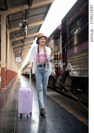 Smiling asian woman holding phone taking photo and dragging purple luggage on train station platform Smiling asian woman holding phone taking photo and dragging purple luggage on train station platform 134621697