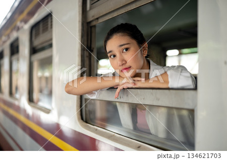 Pretty asian woman leaning head on arms looking out of carriage's window at train station platform. Pretty asian woman leaning head on arms looking out of carriage's window at train station platform. 134621703