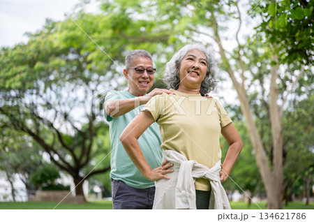 Glasses old man massaging woman shoulders while she standing and smiling on grass lawn in the park. Glasses old man massaging woman shoulders while she standing and smiling on grass lawn in the park. 134621786