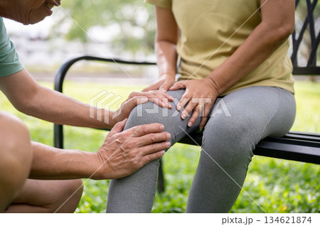 Old man holding or massaging a pained or injury knee of a woman sits on bench after exercise in park 134621874