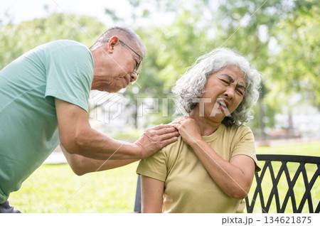 Old man holding or massaging pained or injury shoulder of woman sits on bench after exercise in park 134621875