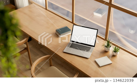 White screen laptop with book and plant on wooden table with chairs in a glass wall cafe or library. 134622039