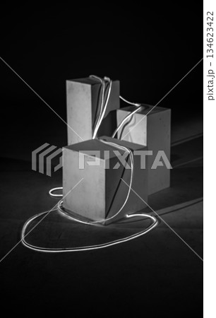 A black-and-white photograph showcases an art installation where geometric blocks, starkly lit, are elegantly bound by a single, glowing cord. A black-and-white photograph showcases an art installation where geometric blocks, starkly lit, are elegantly bound by a single, glowing cord. 134623422