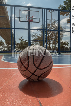 A worn basketball sits on an outdoor court, with a hoop and backboard visible under the Ring Road Bridge in the distance. 134623428