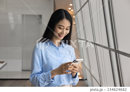 Smiling Asian businesswoman using her smartphone standing in modern office Smiling Asian businesswoman using her smartphone standing in modern office 134624682