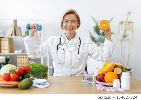 A smiling nutritionist holds a dumbbell and an orange, promoting healthy eating and exercise in her office. A smiling nutritionist holds a dumbbell and an orange, promoting healthy eating and exercise in her office. 134627824