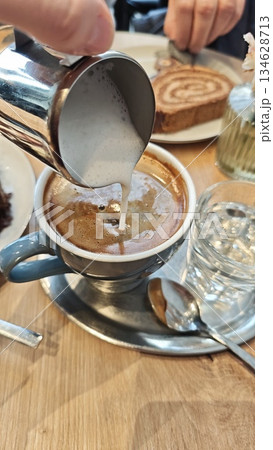Cup of coffee with fresh milk foam being poured from a metal pitcher on a wooden cafe table. The image represents coffee ritual, craftsmanship, and cozy social moment Cup of coffee with fresh milk foam being poured from a metal pitcher on a wooden cafe table. The image represents coffee ritual, craftsmanship, and cozy social moment 134628713