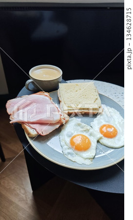 Plate with fried egg ham cheese sandwich and coffee served on a small table in a cozy cafe setting. The image represents classic breakfast routine, comfort food, and relaxed morning lifestyle Plate with fried egg ham cheese sandwich and coffee served on a small table in a cozy cafe setting. The image represents classic breakfast routine, comfort food, and relaxed morning lifestyle 134628715