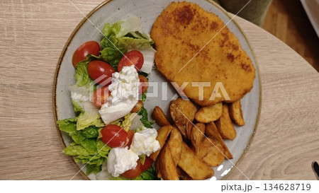 Plate with breaded schnitzel roasted potato and fresh salad with tomato and cheese on a wooden table. The image represents comfort food balanced meal and casual home dining 134628719