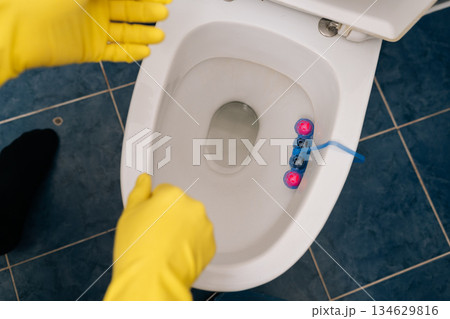 Top view of hands in yellow protective gloves placing toilet freshener block in clean bathroom, highlighting domestic hygiene, daily routine, household cleaning. Concept of hygiene and cleanliness. 134629816