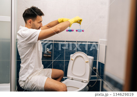 Man wearing yellow rubber gloves placing toilet bowl fresheners in white ceramic toilet, ensuring sanitation and pleasant fragrance in household bathroom. Concept of housekeeping chores and hygiene. 134629819