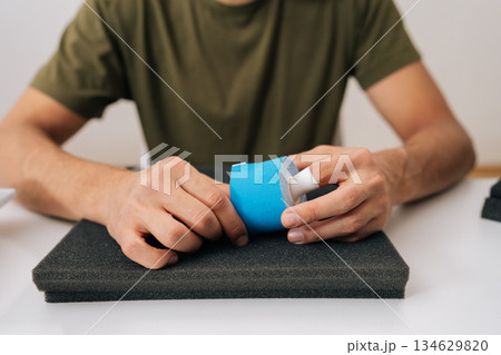 Male hand holding roll of kinesiology tape on table, preparing to apply for muscle, joint or tendon pain relief, representing self-care solution for aches and injuries. 134629820