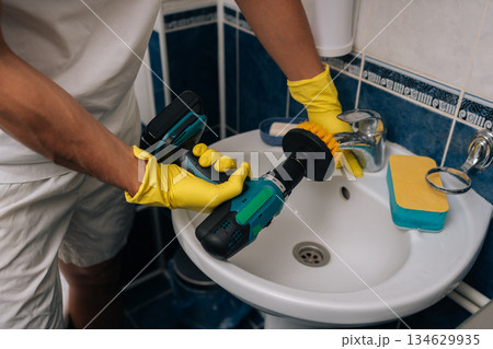 Close-up of man wearing yellow rubber gloves utilizing electric screwdriver with brush attachment for powerful and efficient deep cleaning of bathroom sink and faucet. Concept of hygiene, cleanliness. Close-up of man wearing yellow rubber gloves utilizing electric screwdriver with brush attachment for powerful and efficient deep cleaning of bathroom sink and faucet. Concept of hygiene, cleanliness. 134629935