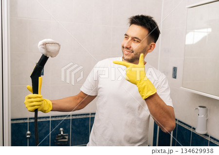 Portrait of man wearing yellow rubber gloves and white t-shirt using steam cleaner in clean bathroom while confidently pointing finger to appliance, showcasing efficient domestic chores and hygiene. Portrait of man wearing yellow rubber gloves and white t-shirt using steam cleaner in clean bathroom while confidently pointing finger to appliance, showcasing efficient domestic chores and hygiene. 134629938
