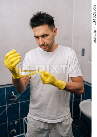 Man frustrated by torn rubber glove, experiencing cleaning problems and household chore difficulties in bathroom, highlighting disappointment with broken equipment and unexpected trouble. Man frustrated by torn rubber glove, experiencing cleaning problems and household chore difficulties in bathroom, highlighting disappointment with broken equipment and unexpected trouble. 134629955