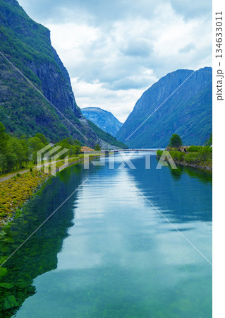 The mouth of the Naeroydalselvi river in the village of Gudvangen on Naeroyfjord on a cloudy day. Reflection of mountains in water. Beautiful nature of Norway. Vertical image 134633011