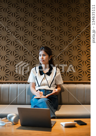 A young woman sits in a modern cafe, holding a notebook, with a laptop nearby, surrounded by stylish decor. A young woman sits in a modern cafe, holding a notebook, with a laptop nearby, surrounded by stylish decor. 134635111