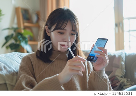 自宅のリビングで体温計を手に持ちスマートフォンでオンライン診療を受ける女性 自宅のリビングで体温計を手に持ちスマートフォンでオンライン診療を受ける女性 134635531