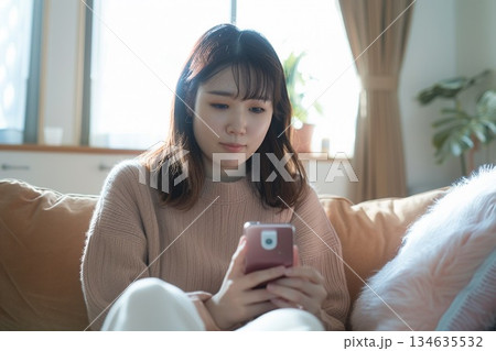 自宅のリビングで体温計を手に持ちスマートフォンでオンライン診療を受ける女性 134635532