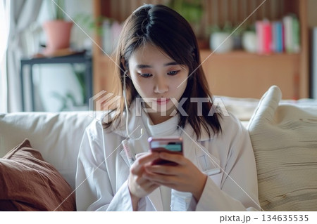 自宅のリビングで体温計を手に持ちスマートフォンでオンライン診療を受ける女性 134635535