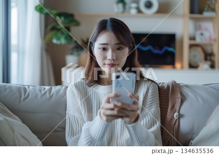 自宅のリビングで体温計を手に持ちスマートフォンでオンライン診療を受ける女性 134635536