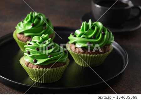 Three chocolate cupcakes with green whipped cream and chocolate sprinkles on dark brown background. St. Patrick's Day. Close up. 134635669