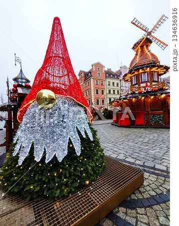 Large decorative gnome figure, with a red, cone-shaped hat, silver beard, and a spherical gold nose, crafted from evergreen branches with lights and wooden Christmas stall topped with windmill at 134636916