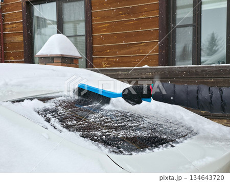 Woman clearing snow from her car windshield during winter. Preparing vehicle for travel in cold weather. Winter season car maintenance. 134643720