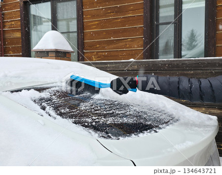 Woman clearing snow from her car windshield during winter. Preparing vehicle for travel in cold weather. Winter season car maintenance. Woman clearing snow from her car windshield during winter. Preparing vehicle for travel in cold weather. Winter season car maintenance. 134643721